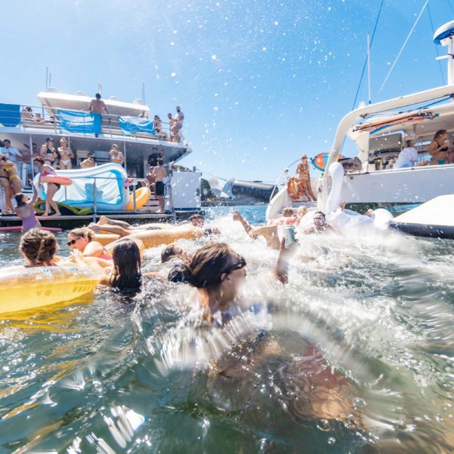 A group of people swimming and splashing in water near boats on a sunny day. The scene includes individuals on inflatable tubes, other swimmers, and boats with people on board, one of which could be a luxury yacht hire Sydney.
