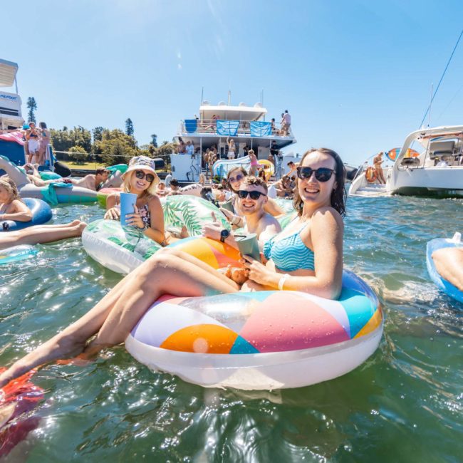A group of people enjoying a sunny day on the water with flotation devices near several anchored boats, making the most of their luxury yacht hire in Sydney.