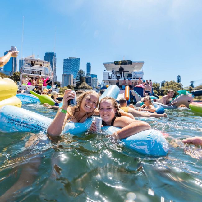 Two people float on inflatable devices holding drinks in a crowded water area with other people and boats nearby. Skyscrapers are visible in the background on a sunny day, perfect for a catamaran party Sydney.