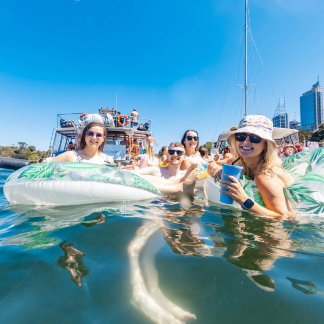 Four people are sitting on inflatable loungers in the water, holding drinks and smiling. Boats and other people enjoying a Sydney boat party hire are visible in the background, with a cityscape in the distance.