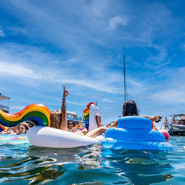 People float on inflatable toys in a body of water on a sunny day with boats and a clear sky in the background, enjoying the vibrant atmosphere of a Luxury yacht hire Sydney.
