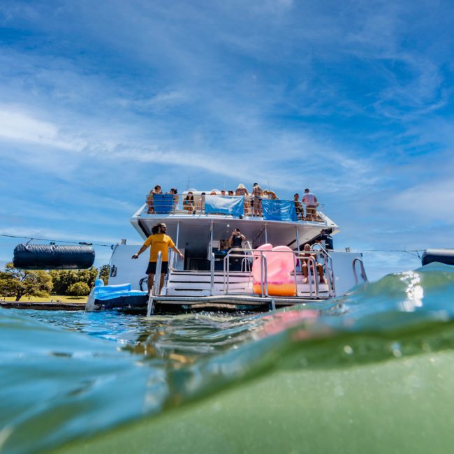 A boat with people on deck is anchored in clear, shallow water under a blue sky. The lower part of the image is taken from a perspective half-submerged in water, capturing the essence of a private yacht charter Sydney Harbour.