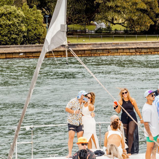 A group of people stand and sit on a boat on the water. Trees and a grassy area can be seen in the background, making it an ideal scene for Sydney boat party hire.