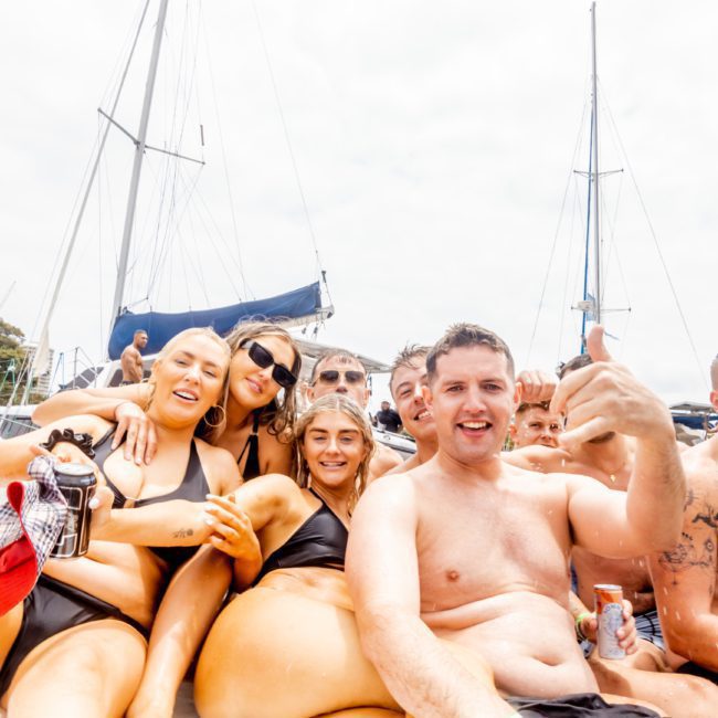 A group of eight people in swimwear are sitting together on a boat, smiling and posing for the camera at a catamaran party in Sydney. They appear to be enjoying a sunny day with boats and a cityscape in the background.