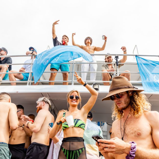 A group of people enjoy a lively and festive Catamaran party Sydney on a boat. Some individuals are dancing, holding drinks, and using their phones, while others observe from the upper deck. The atmosphere is vibrant and fun.