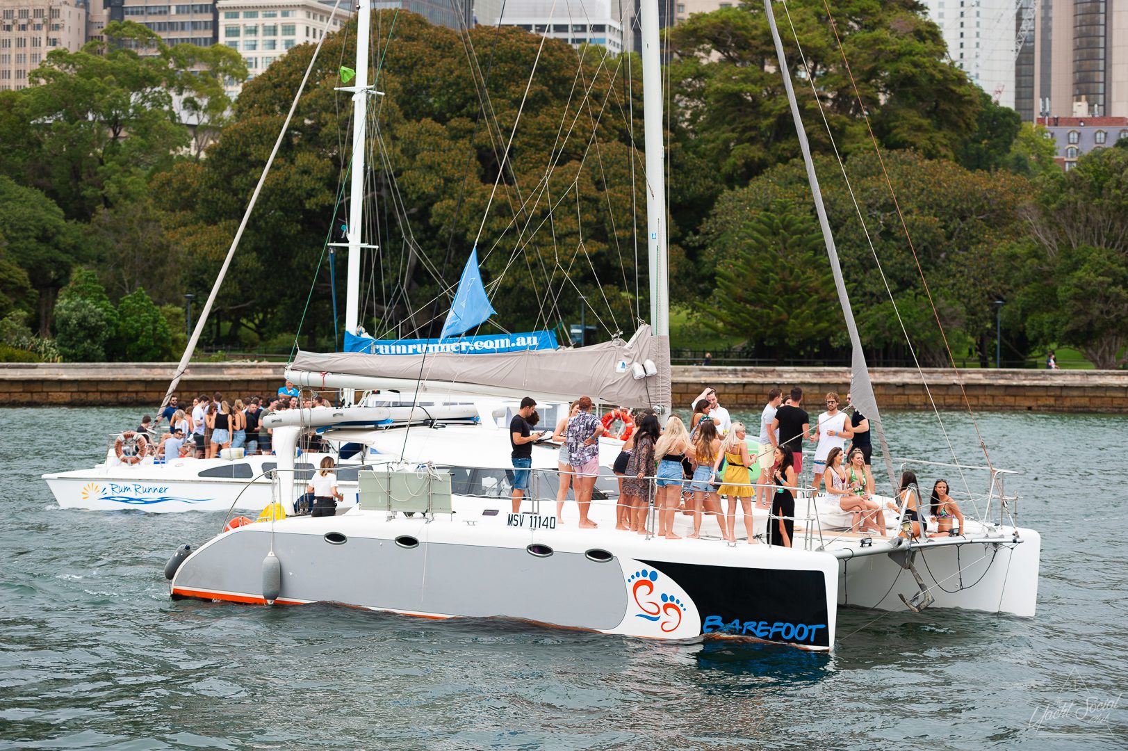 A large catamaran is full of people, dressed casually, enjoying a sunny day on a river with trees and city buildings in the background. It's the perfect setting for a Catamaran party Sydney, creating unforgettable moments on the water.