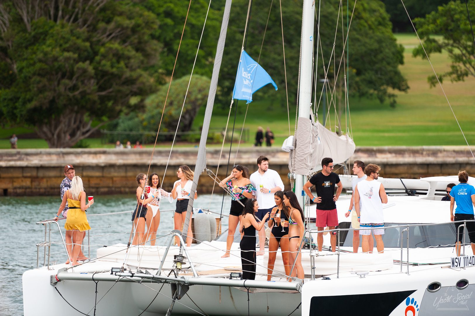 A group of people stands and socializes on a docked luxury yacht near a green park or wooded area. Some carry drinks, and they appear to be enjoying a casual Corporate boat event in Sydney.