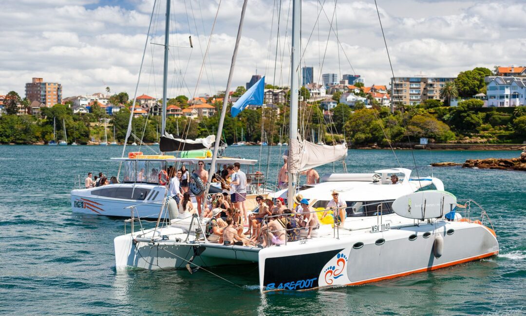 People are gathered on the deck of two large sailboats in a calm body of water, with a cityscape and green shoreline in the background. Perfect for those looking to experience luxury yacht hire Sydney or plan a Catamaran party Sydney. The sky is partly cloudy.