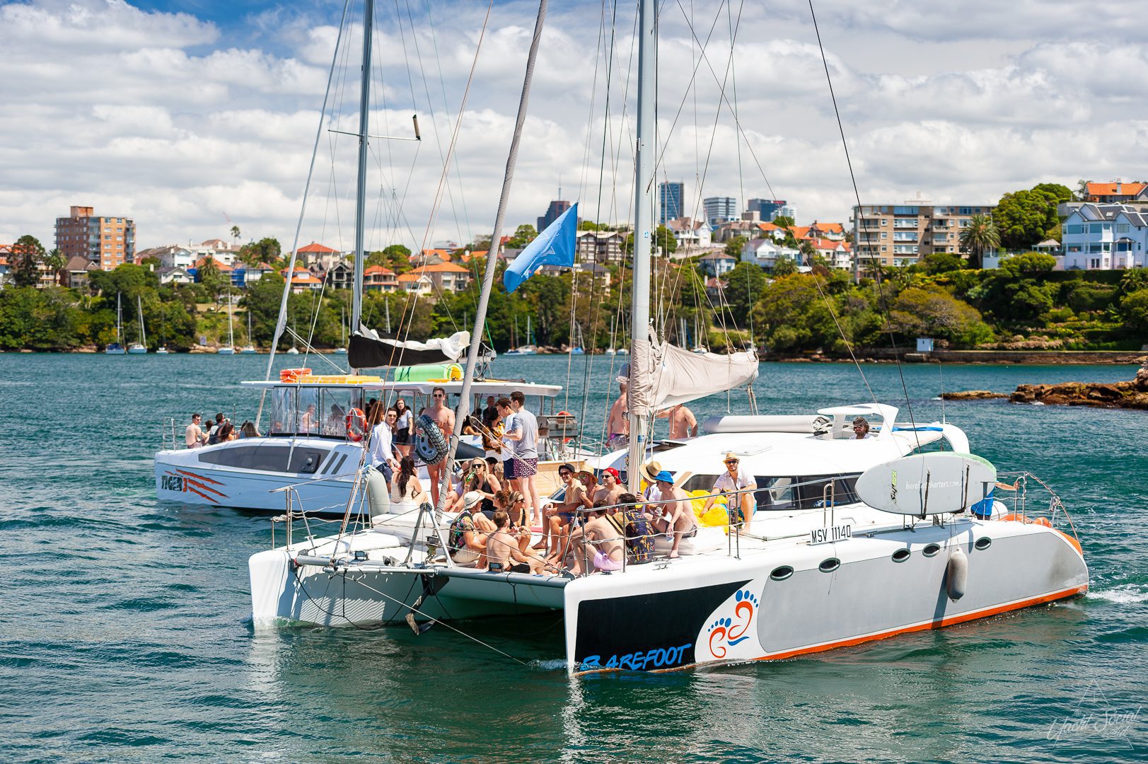 People are gathered on the deck of two large sailboats in a calm body of water, with a cityscape and green shoreline in the background. Perfect for those looking to experience luxury yacht hire Sydney or plan a Catamaran party Sydney. The sky is partly cloudy.