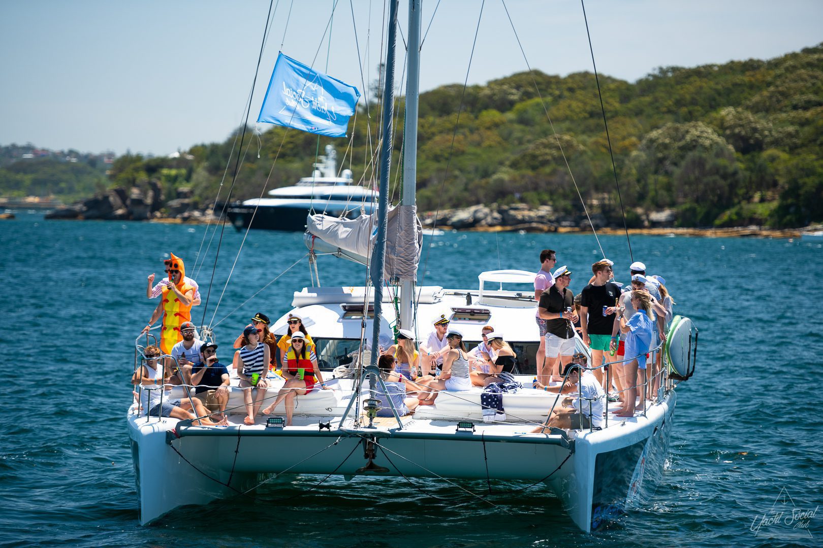 A group of people enjoying a sunny day on a large catamaran in a scenic body of water, with lush greenery and other boats in the background. Some participants are in costumes or casual attire, perfect for a lively Catamaran party Sydney.