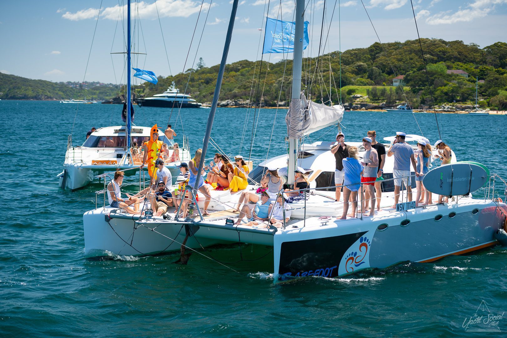 A group of people is gathered on a white catamaran in a scenic bay, surrounded by clear blue water with a tree-covered shoreline in the background. Perfect for corporate boat events Sydney, this luxury yacht hire offers an unforgettable experience amidst breathtaking views.