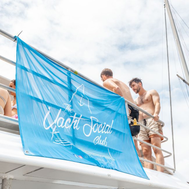 A blue banner with "The Yacht Social Club" printed on it hangs on a yacht railing. Shirtless people are socializing on the deck, enjoying the vibrant atmosphere of the Catamaran party in Sydney.