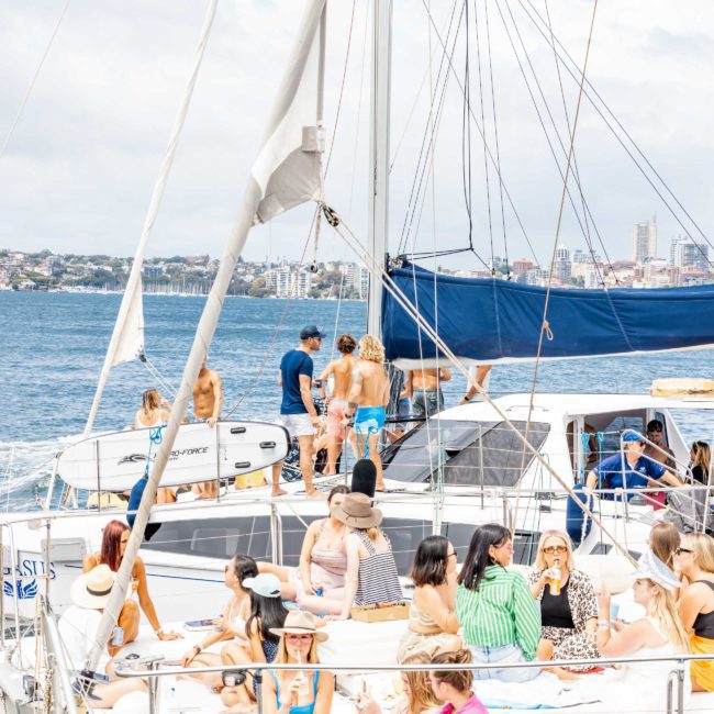 A group of people socializing on a luxury yacht hire Sydney in open water, with a cityscape visible in the background. Some people are seated, while others stand talking on the deck.