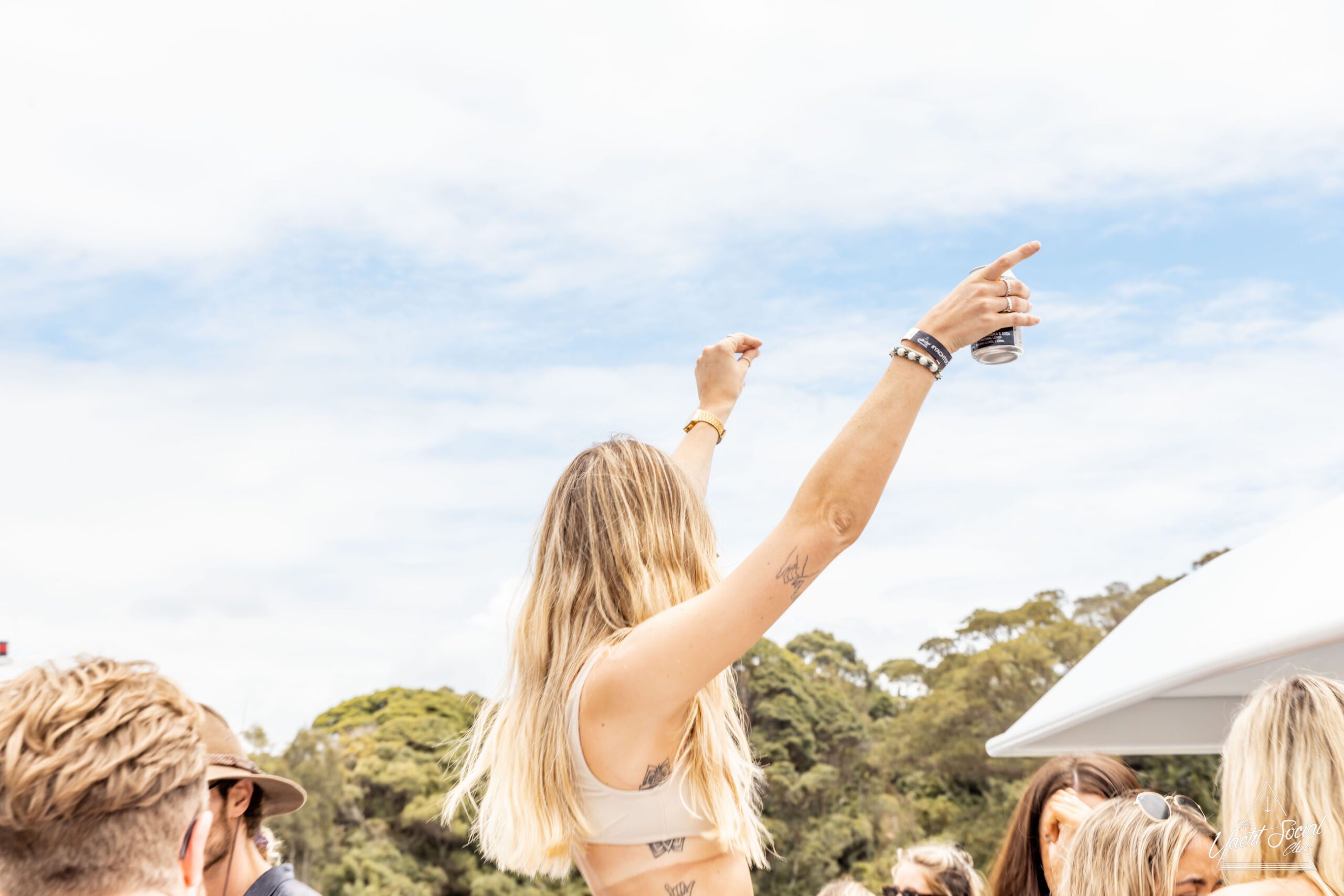 A blonde woman raises her arms in the air while holding a drink, surrounded by other people outdoors on a sunny day at a lively Sydney boat party hire.