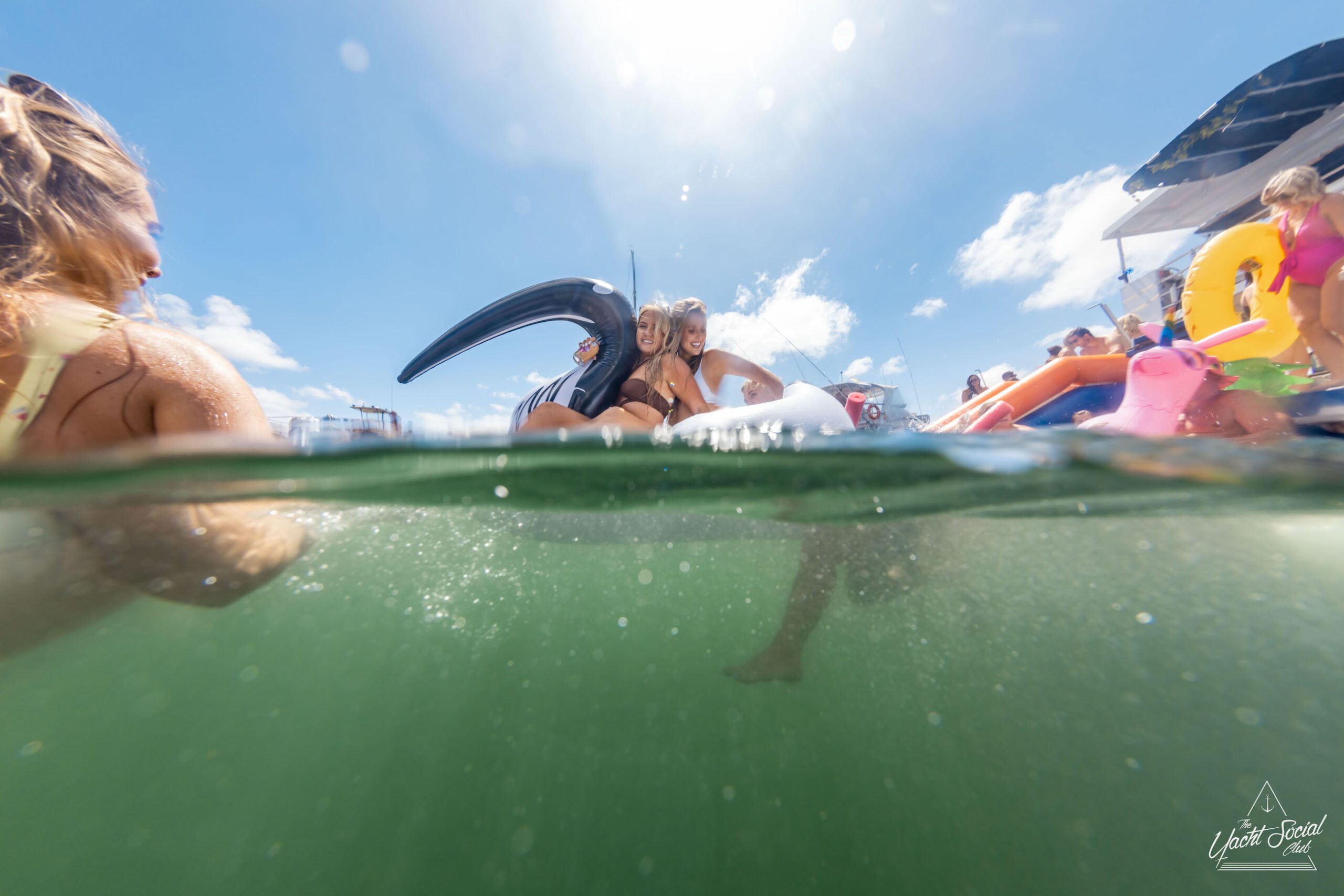 People are enjoying a sunny day at the beach, with some floating on inflatable toys in the water while others indulge in a Sydney boat party hire. The photo is partially submerged, showing both above and below the water's surface.