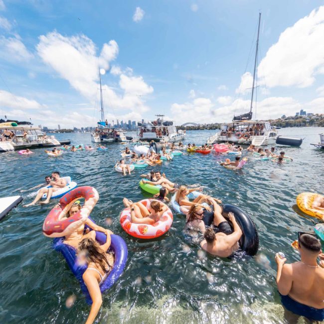 People on inflatables in the water with boats nearby on a sunny day. Some individuals are swimming while others relax on the inflatables, enjoying a Sydney boat party hire, with the background featuring a distant city skyline.