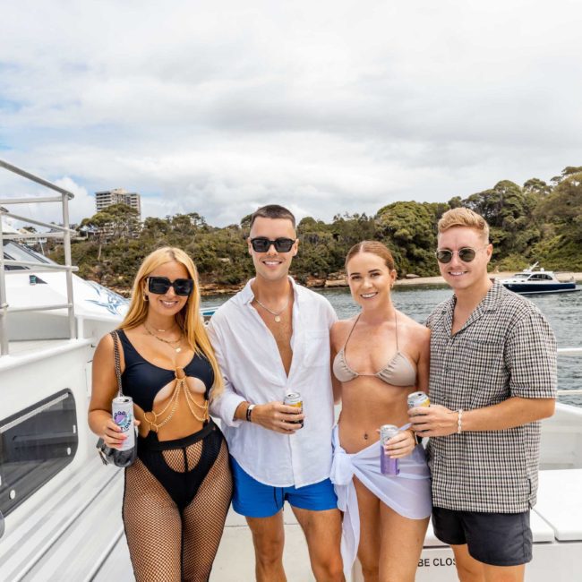 Four people in swimwear, holding drinks, stand on a boat with water and trees in the background. Experience the ultimate catamaran party Sydney, with friends and stunning views.