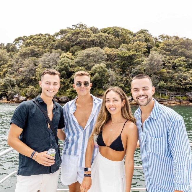 Four people are standing on the deck of a boat, smiling at the camera. Trees and water are visible in the background during a private yacht charter Sydney Harbour.