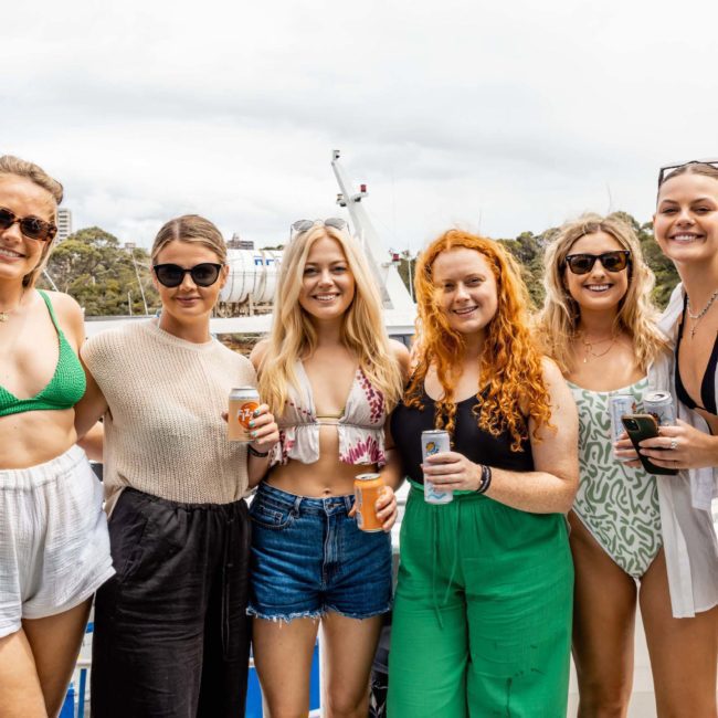 Six women smiling and holding drinks, standing together on a catamaran with water and trees in the background. They are dressed in casual summer attire, enjoying a Catamaran party in Sydney.