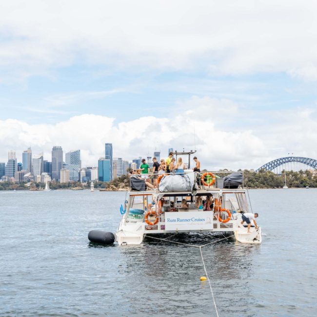 A boat named "Rum Runner Cruises" is anchored in a bay with several people on board, enjoying the view of the city's skyline and a bridge under a cloudy sky. For an unforgettable experience, consider DJ boat hire Sydney or Catamaran party Sydney for your next adventure.