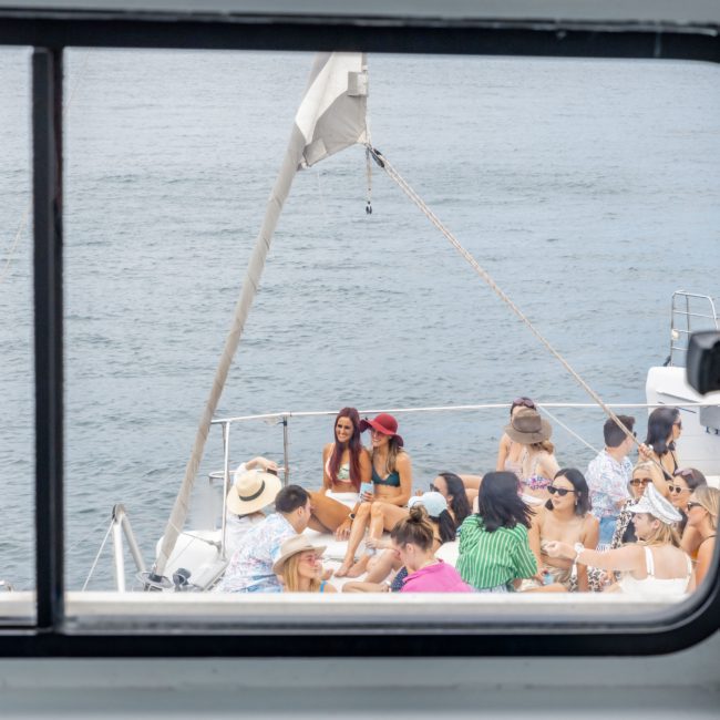 A group of people relaxes on the deck of a private yacht charter on a calm body of water, viewed through a window frame.