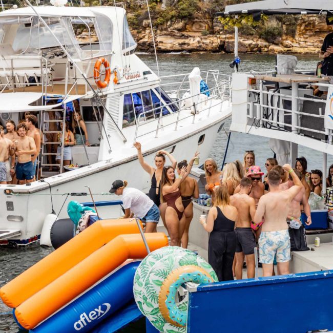 A group of people are on boats and a floating dock, some engaging with an inflatable slide and water toys. The scene includes music equipment and appears to be a lively Sydney boat party hire in a coastal area.