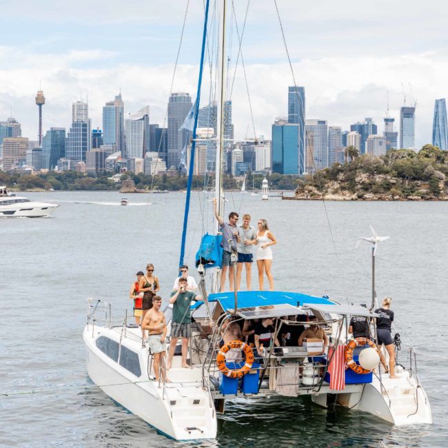 A group of people are on a sailboat in a harbor with the stunning Sydney skyline in the background. Some are standing on the boat while others are seated or standing near the railings, enjoying what looks like a delightful Sydney boat party hire experience.