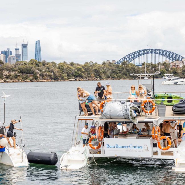 Two boats docked side by side on the water with passengers on board. Skyline and Sydney Harbour Bridge visible in the background. The larger boat displays a "Rum Runner Cruises" sign. Enjoy private yacht charter Sydney Harbour or corporate boat events in this stunning setting.