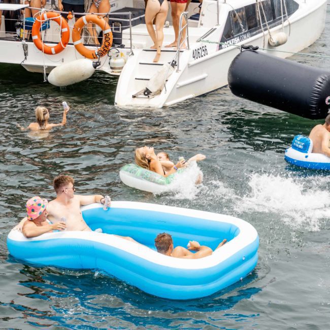 People enjoying a summer day on the water, some swimming, some using floats, and others on boats. Luxurious private yacht charters in Sydney Harbour are making the day even more special.