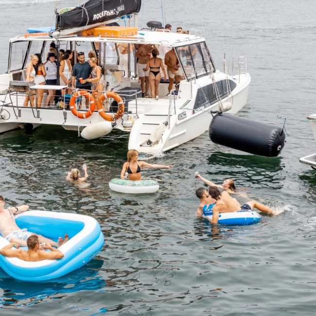 A group of people enjoying a day on the water with boats and inflatable pools. Some are on the boats while others relax and swim in the water, embodying the perfect Sydney boat party hire experience.