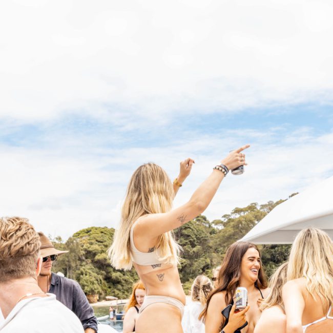 Group of people enjoying a sunny day on a boat, with one person standing and pointing, others smiling and talking. Trees are visible in the background. Perfect for a catamaran party in Sydney or a private yacht charter on Sydney Harbour.