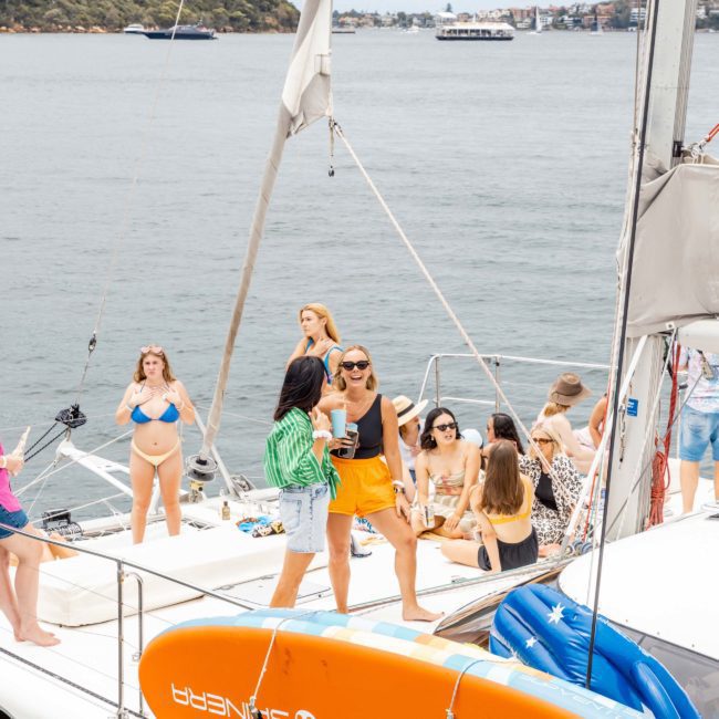 A group of people, some in swimwear, are gathered on the deck of a sailboat, socializing and enjoying the view. An orange surfboard labeled "SUNSEEKER" is mounted on the boat. The scene hints at a lively DJ boat hire Sydney event or even a fun catamaran party Sydney experience.
