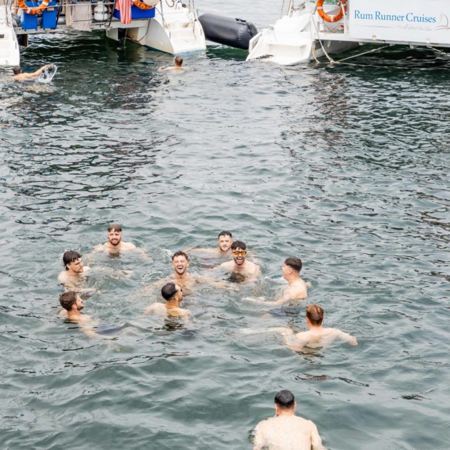 A group of people swim and float in the water between two anchored boats. The boats have banners, one of which reads "Rum Runner Cruises." For an unforgettable experience, consider DJ boat hire Sydney for your next outing or a Catamaran party Sydney to elevate your fun.