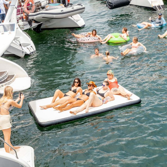 People are relaxing on inflatable mattresses and floating devices in the water near several boats. A woman is taking a photo of three women on a large floating mat, capturing memories of their Private yacht charter Sydney Harbour experience.