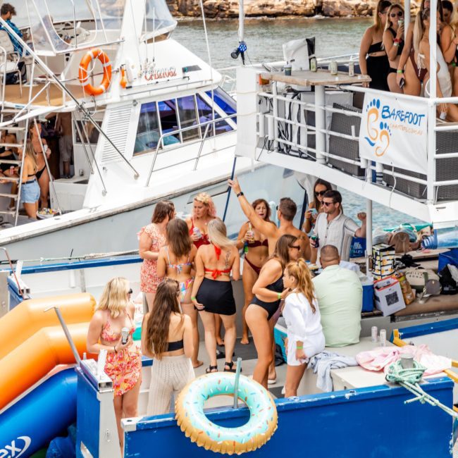 A group of people in swimwear are socializing and dancing on a boat equipped with slides. Another boat with additional people is docked nearby, highlighting the fun atmosphere of a Sydney boat party hire. The scene suggests a laid-back outdoor boat party.