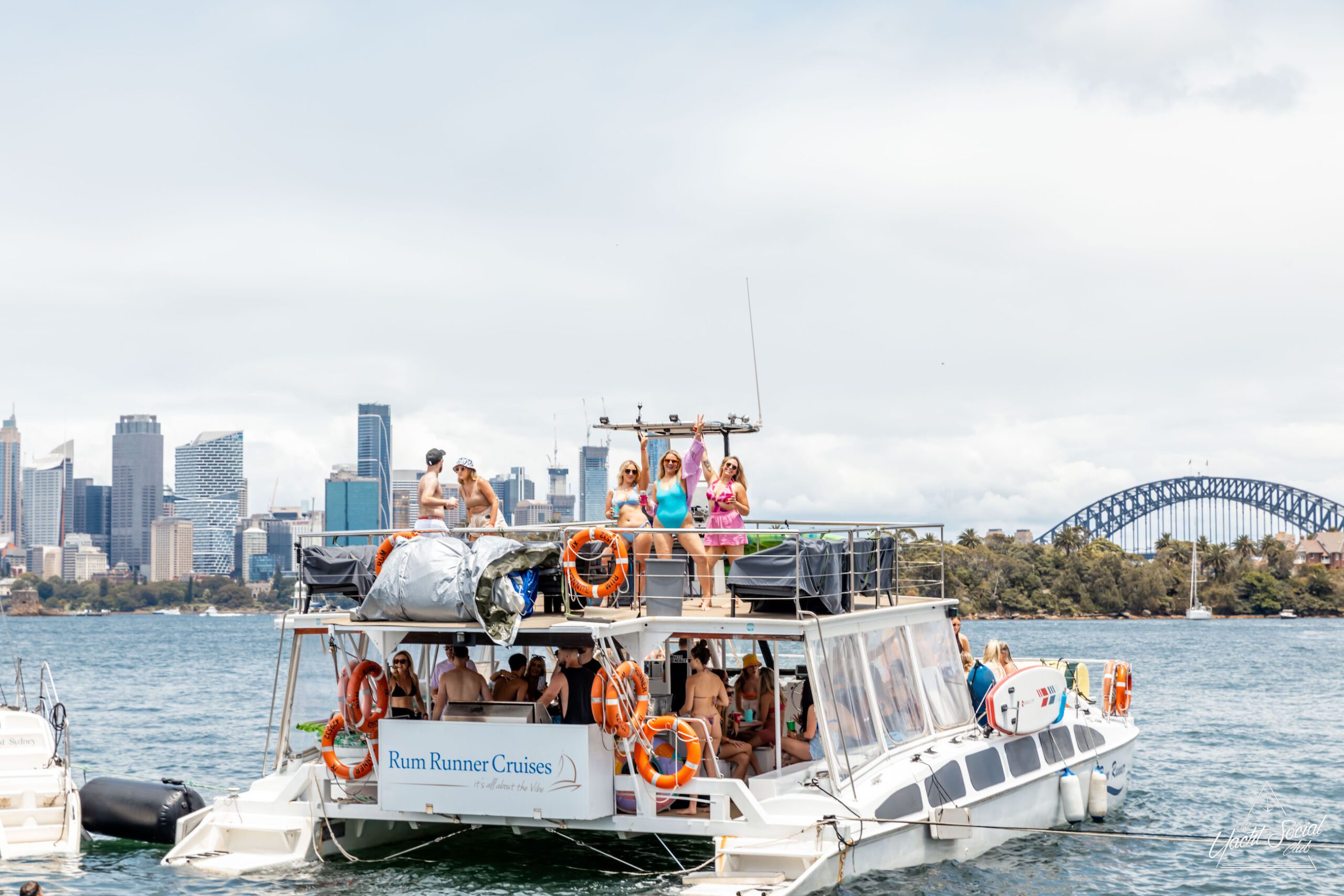 A luxury yacht named "Rum Runner Cruises" with people on board is sailing on the water. A city skyline and a bridge are visible in the background, offering a stunning view of Sydney.
