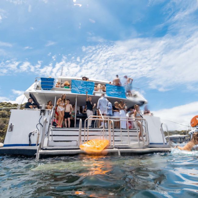 A group of people are gathered on a luxury yacht hire Sydney in open water under a partly cloudy sky. Some are standing and others are seated, while one person swims nearby. A sailboat is in the background, creating the perfect setting for a memorable Sydney boat party hire.
