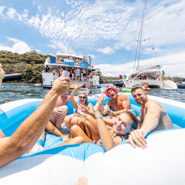 A group of people in swimsuits enjoy themselves on an inflatable raft on the water, with several boats and other people in the background. Nearby, a catamaran party Sydney adds to the vibrant atmosphere.