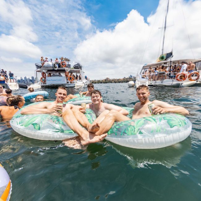 Three men relax on inflatable tubes in the water, surrounded by boats with people on board. The sky is partly cloudy, suggesting a sunny day suitable for water activities, perhaps even a luxury yacht hire Sydney experience.