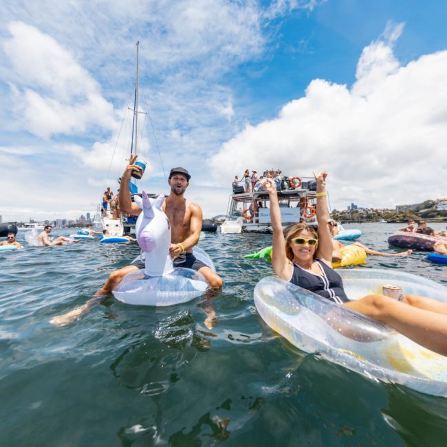 A group of people enjoy a sunny day on the water, floating on inflatable rings with boats in the background. Some are raising their arms and drinks in celebration, while others discuss their upcoming DJ boat hire Sydney event.