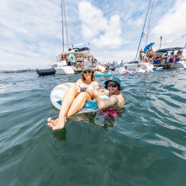 Two people are floating on an inflatable ring in the water, with several boats and other people in the background under a partly cloudy sky. A private yacht charter in Sydney Harbour adds a touch of luxury to the scene.