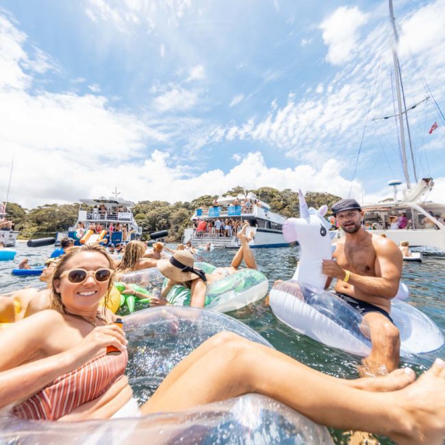 A group of people relax on inflatable floats in a sunny lake, surrounded by elegant boats during a private yacht charter Sydney Harbour.