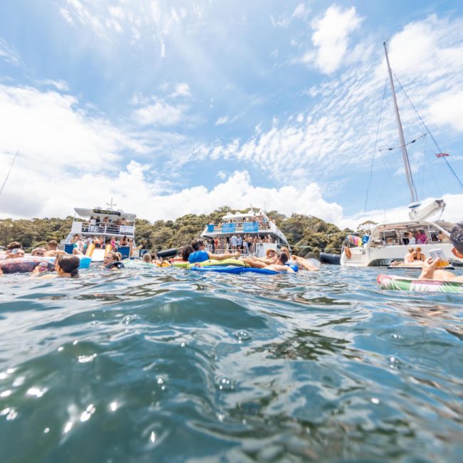 People in swimsuits are floating in the water near boats on a sunny day with clear skies. Some are on inflatable rafts, while others are swimming. The background has trees and more boats, perfect for a luxury yacht hire Sydney adventure.