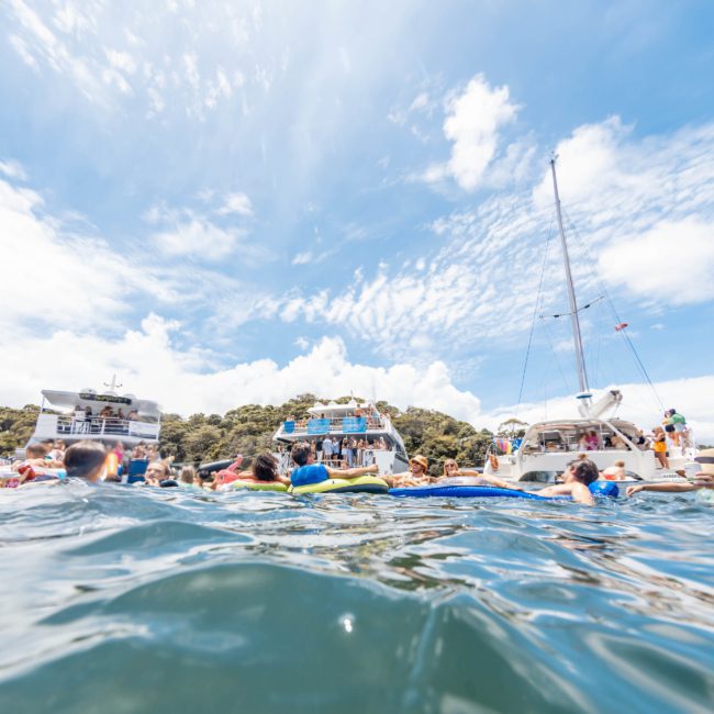 People swimming in the ocean with boats, including a luxury yacht hire in Sydney Harbour, in the background under a partly cloudy sky.