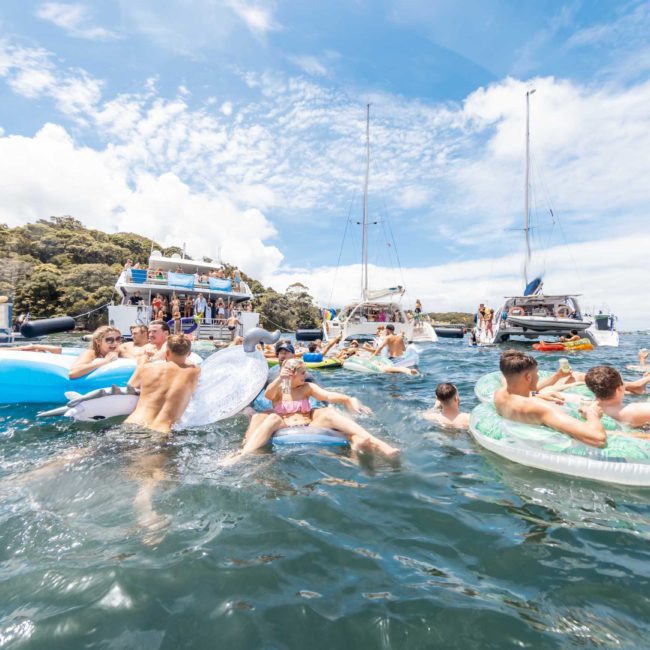A large group of people enjoying a sunny day on the water, with some on inflatable floats and others on boats. The scene includes clear blue skies, lush greenery in the background, and multiple anchored boats, perfectly capturing the essence of a private yacht charter Sydney Harbour.