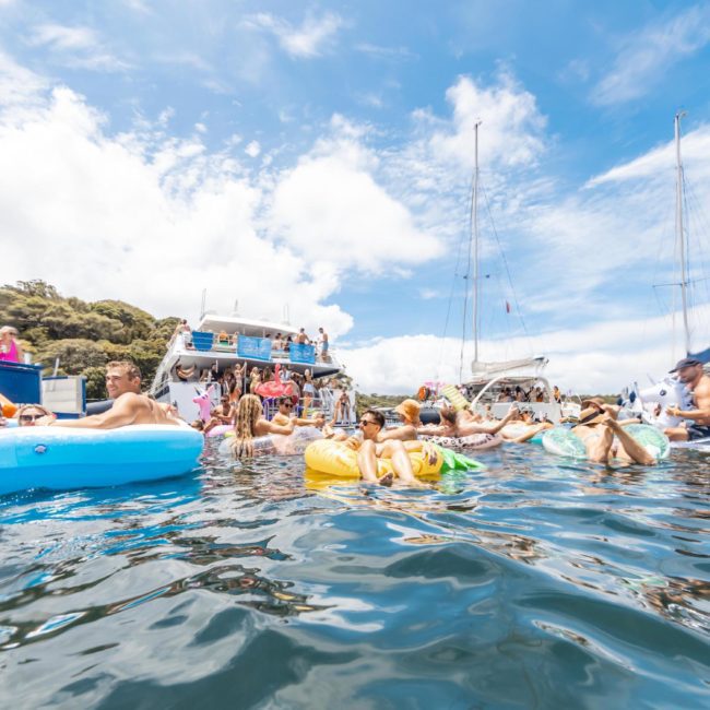People are enjoying various water activities near boats on a sunny day, with some on inflatable floaties and others swimming in the water. Several boats, including a luxury yacht hire Sydney, and a lush hill are visible in the background.