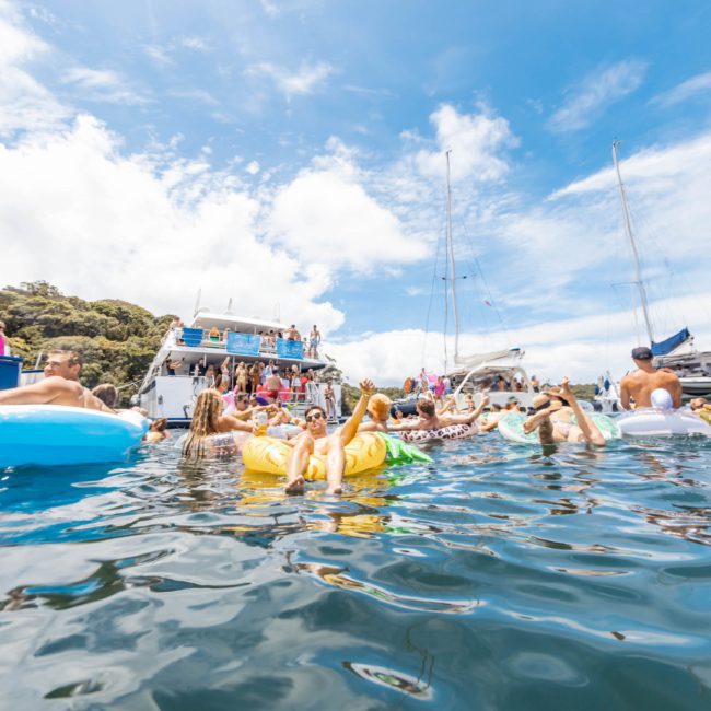 People floating on inflatables in a waterway near anchored boats on a sunny day under a blue sky with scattered clouds, enjoying the fun of a DJ boat hire Sydney.