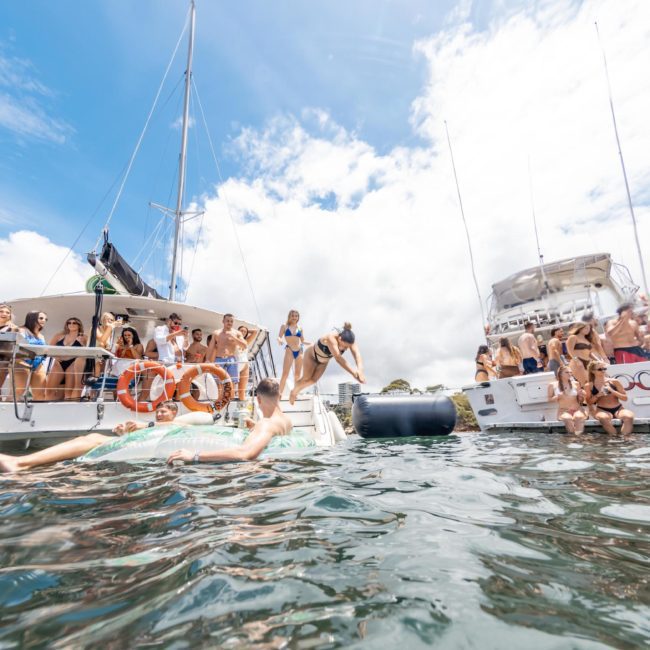People are relaxing and socializing on two boats anchored close to each other in the water, with some individuals swimming and using floats. Enjoy the experience with a luxury yacht hire in Sydney, making your day even more special.