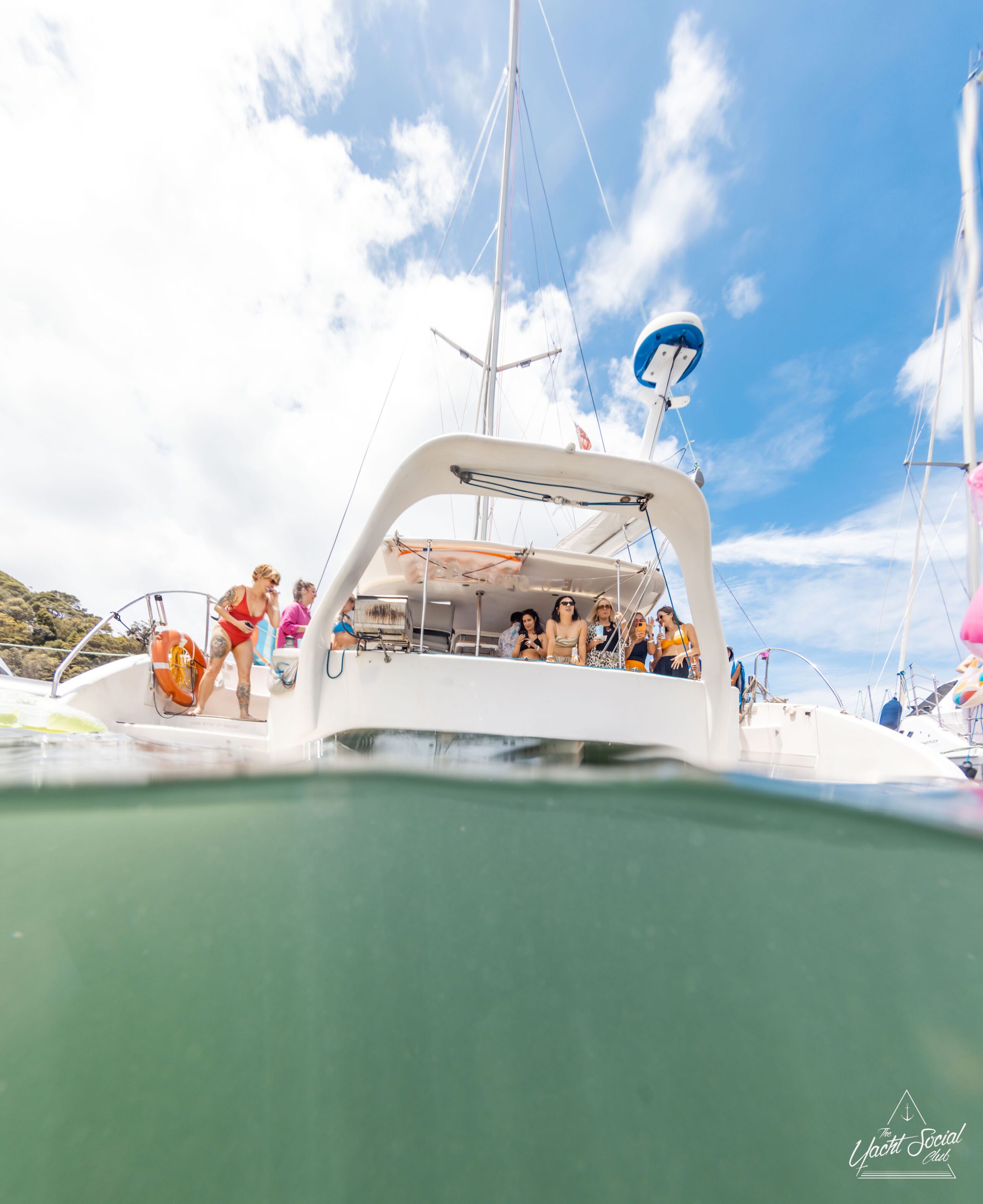 A group of people are enjoying leisure time on a luxury yacht hire in Sydney, with some on deck and others swimming in the water. The sky is partly cloudy.