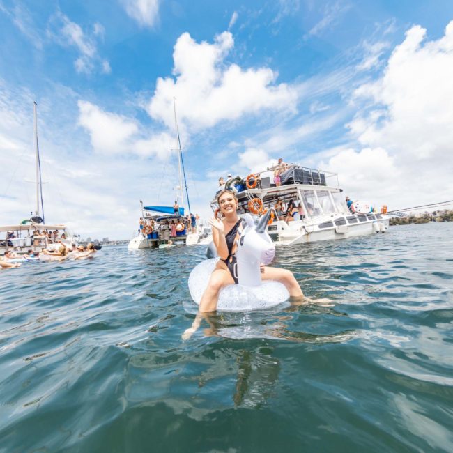 A person rides an inflatable float in the water with boats and more people in the background under a partly cloudy sky, enjoying a private yacht charter Sydney Harbour.